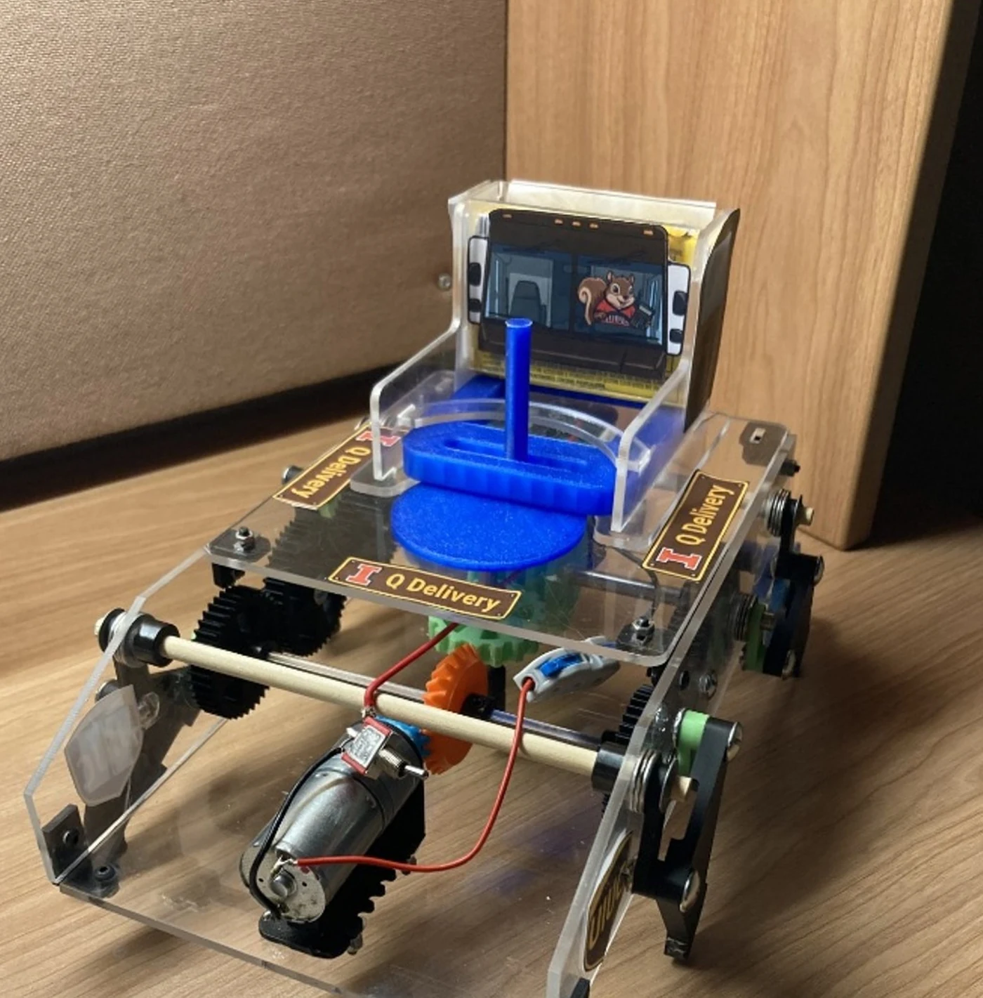The completed ME 370 dispensing walker: a clear acrylic four-legged chassis with the blue Scotch-yoke dispenser and gum hopper on top, driven by a single motor through the gear train below.